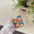 Hand holding a folded retro striped Samsung Z Flip case with a multi-color crochet smiling flower hand strap.
