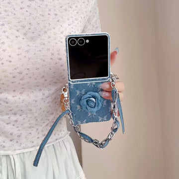 Hand holding an unfolded Samsung Galaxy Z Flip case featuring a 3D denim flower and hanging wrist chain strap.