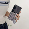Hand holding open Samsung Galaxy Z Flip in a charcoal gray paisley tapestry case with attached heart mirror charm.