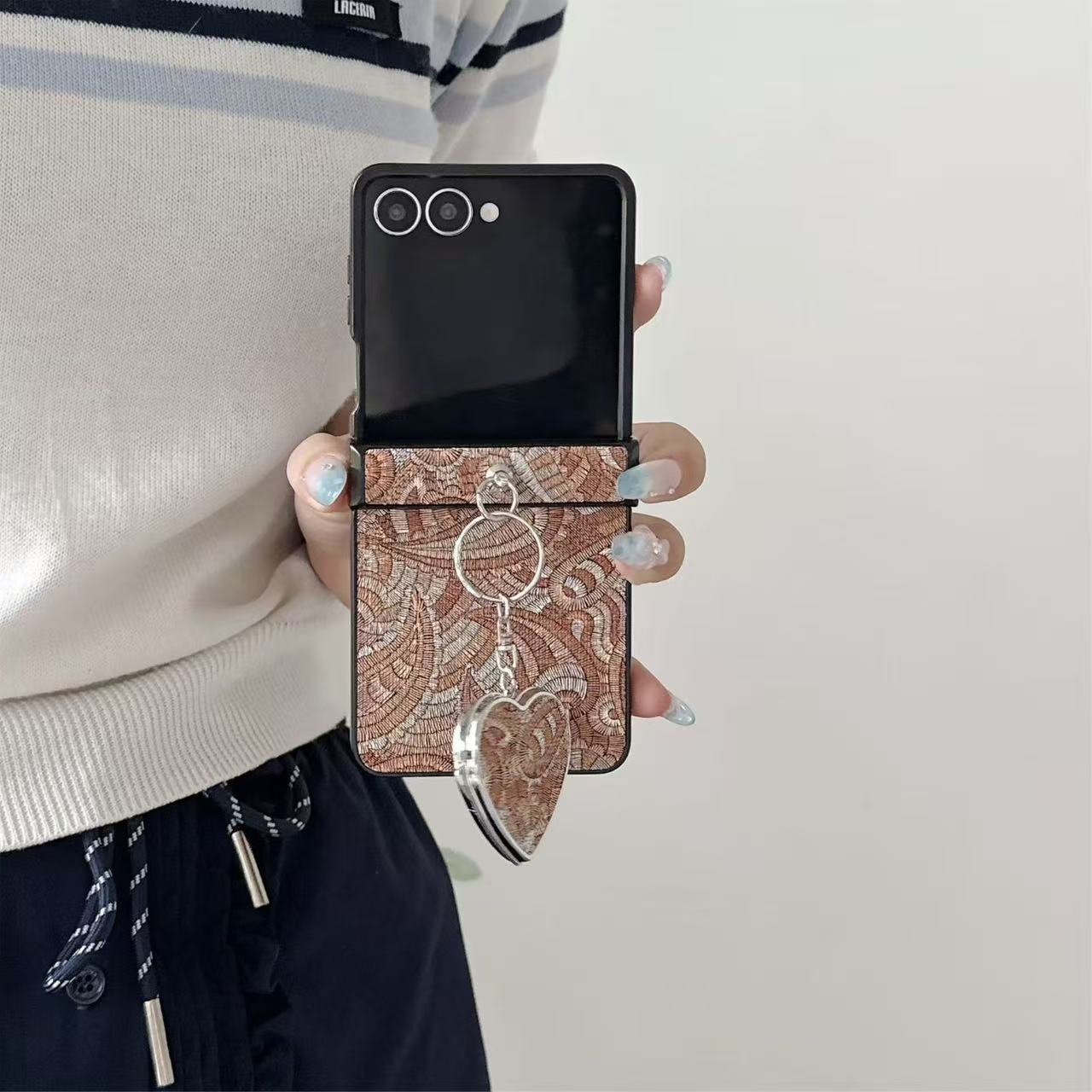 Hand holding unfolded Samsung Galaxy Z Flip phone in an earthy brown woven paisley case with heart mirror keychain attached.