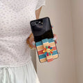 Lifestyle view of a person holding an open Galaxy Z Flip case with vibrant horizontal stripes and smiley flower grip.⁠