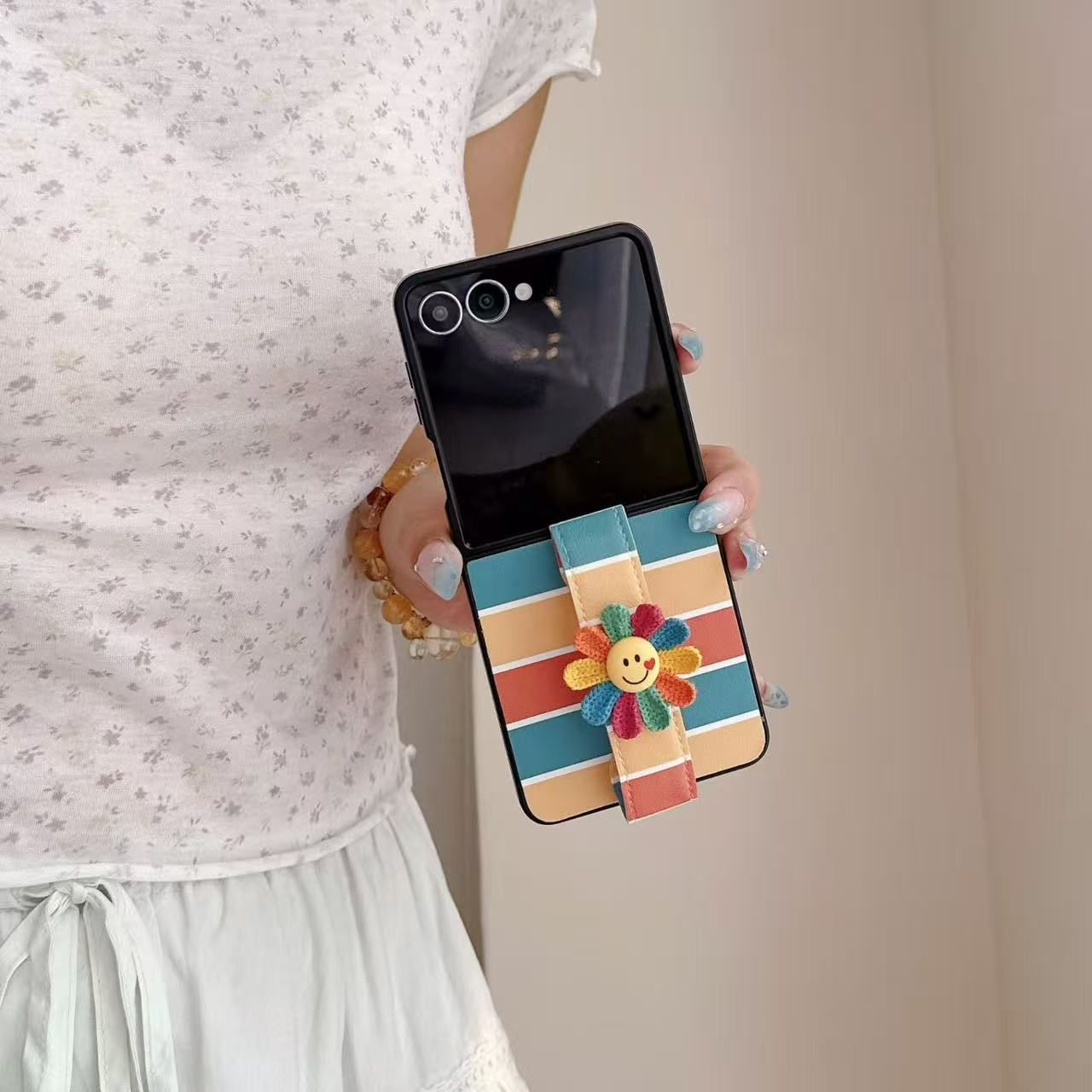 Lifestyle view of a person holding an open Galaxy Z Flip case with vibrant horizontal stripes and smiley flower grip.⁠