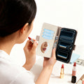 Light-brown pebbled leather smartphone flip case. The large upper panel shows a woman (seen from behind) applying lipstick using the internal oval vanity mirror of the opened case.