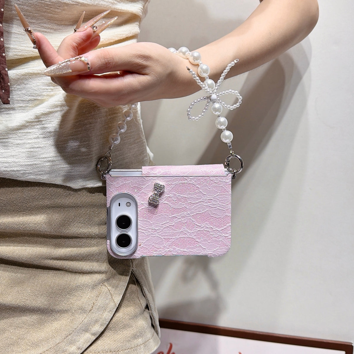 Light pink lace case for iPhone DUO held in hand showing front lock screen display, floral glitter lace back texture, crystal bow charm, and attached pearl bow lanyard chain strap.