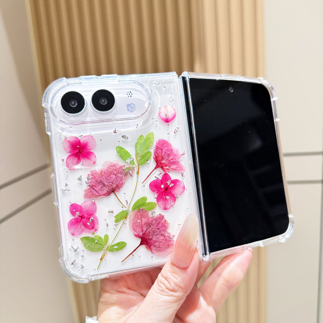 Close-up vertical perspective of a hand holding the clear iPhone DUO real pressed pink flower case, highlighting silver foil flecks and detailed leaf stems.