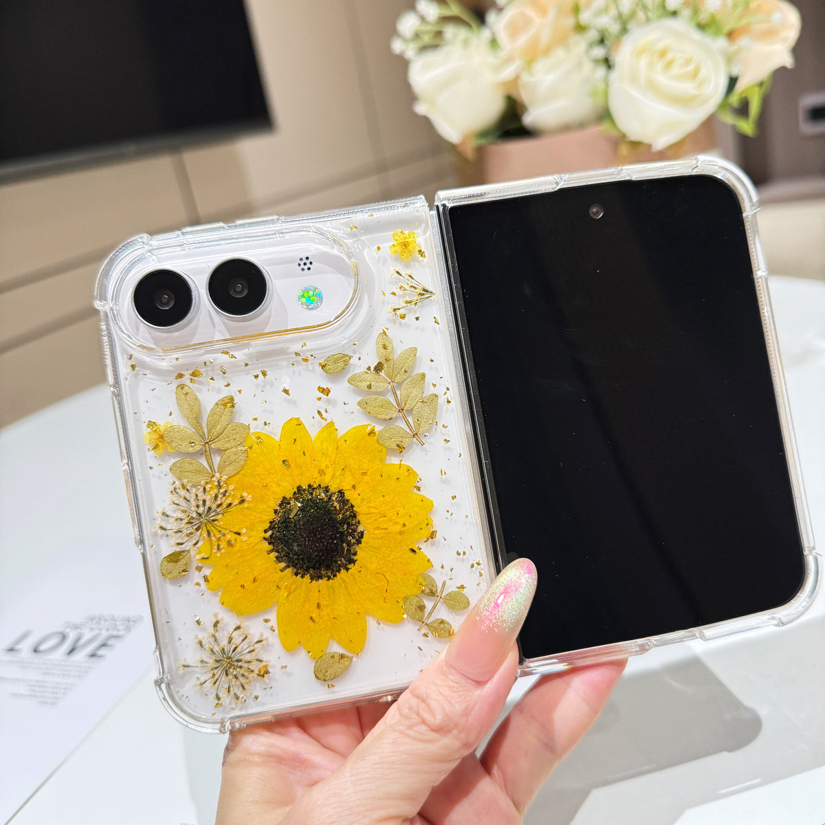 Open view of a hand holding the clear iPhone DUO case showcasing the full real pressed sunflower and botanical arrangement alongside the active display screen.