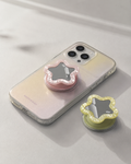 Pastel pink and creamy yellow star-shaped mirror phone grip shown on iPhone with clear case — compact vanity mirror pop socket, flat lay on white surface

