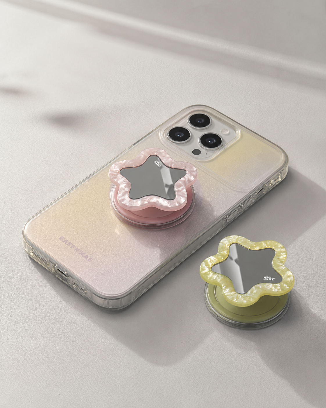 Pastel pink and creamy yellow star-shaped mirror phone grip shown on iPhone with clear case — compact vanity mirror pop socket, flat lay on white surface

