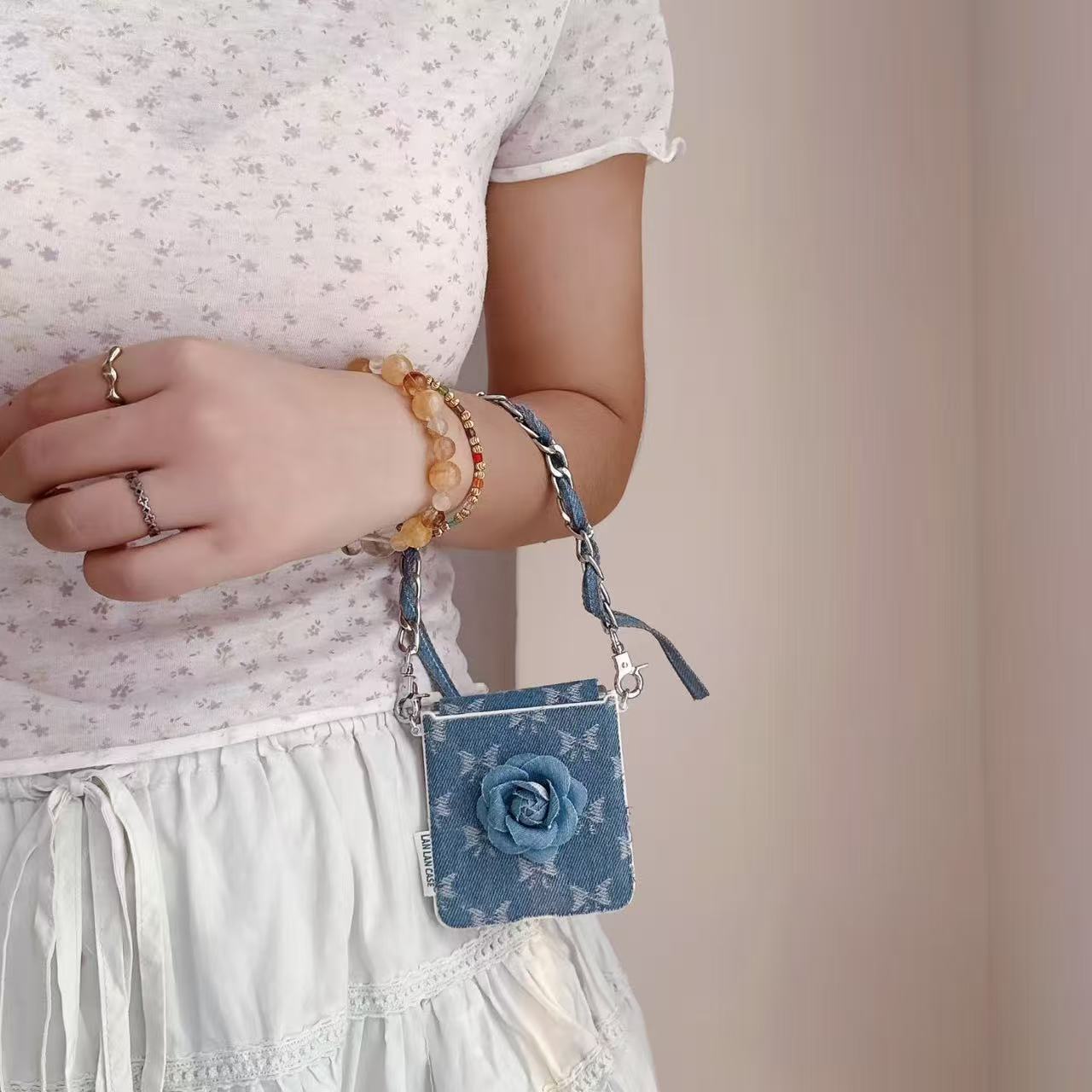 Person wearing light blue skirt holding folded Samsung Z Flip denim flower case by its wrist lanyard.