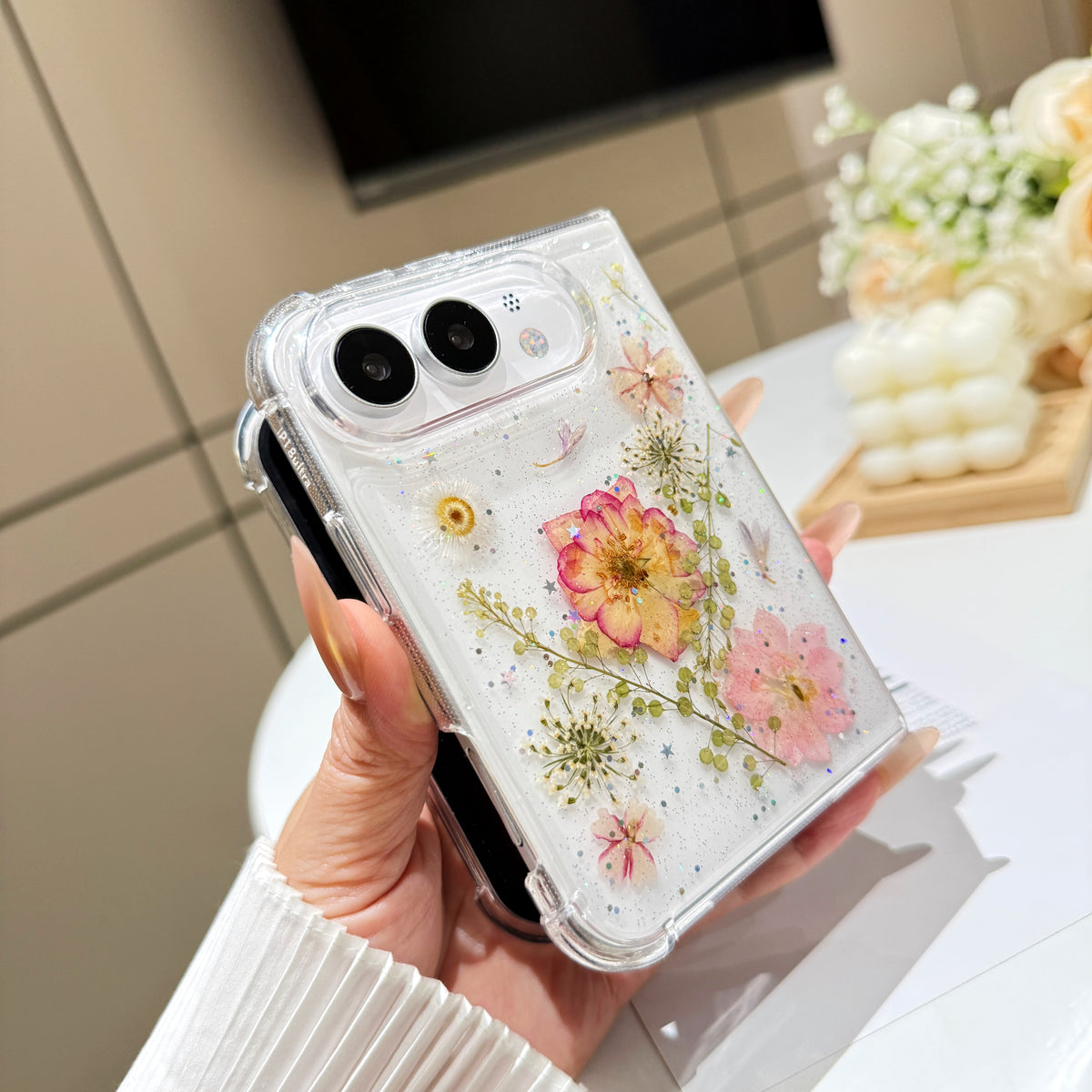 Perspective view of a hand holding the clear pressed flower and star glitter case for iPhone DUO, showing raised camera bezel protection and corner bumper frame.