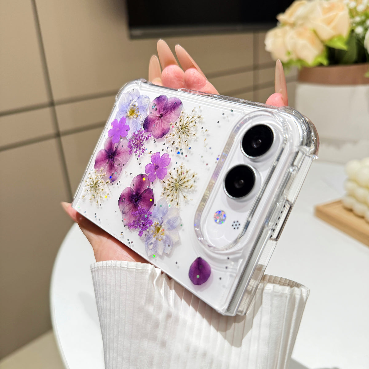 Perspective view of a hand holding the clear purple pressed flower case for iPhone DUO, showing raised camera bezel protection and corner bumper frame