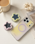 Pink and creamy yellow star mirror phone grips popped out as kickstand next to MagSafe compatible iPhone — expandable phone grip with built-in mirror, aesthetic desk setup

