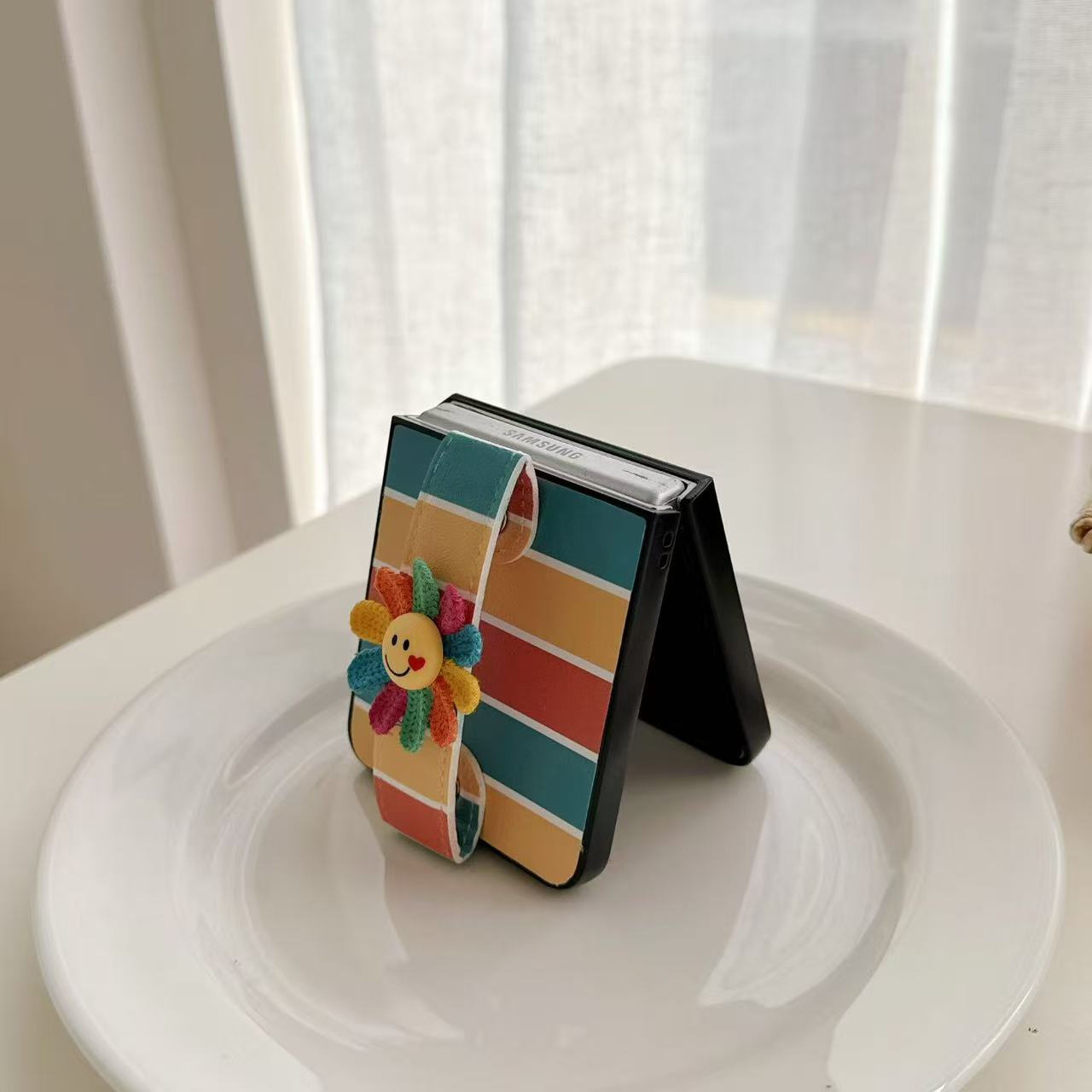 Samsung Galaxy Z Flip standing half-open on a ceramic plate showcasing the back strap and flower charm.