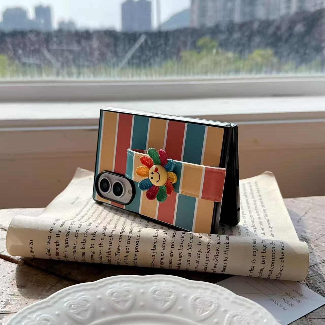 Samsung Galaxy Z Fold in folded standing position resting on a newspaper using the rainbow stripe leather case and flower kickstand strap