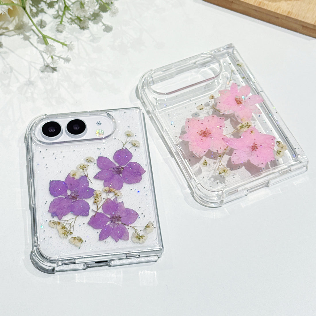 Side-by-side display of two iPhone DUO clear glitter cases showcasing the handmade real pressed flower options in elegant violet purple and soft blossom pink.