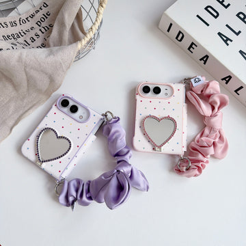 Side-by-side view of lavender purple and soft pink iPhone DUO polka dot cases with rhinestone heart mirrors and matching silk scrunchie wristlet straps.