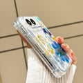 Side perspective view of a hand holding the clear blue blossom pressed flower case for iPhone DUO, showing raised camera bezel protection, corner bumper frame, and side button alignment.