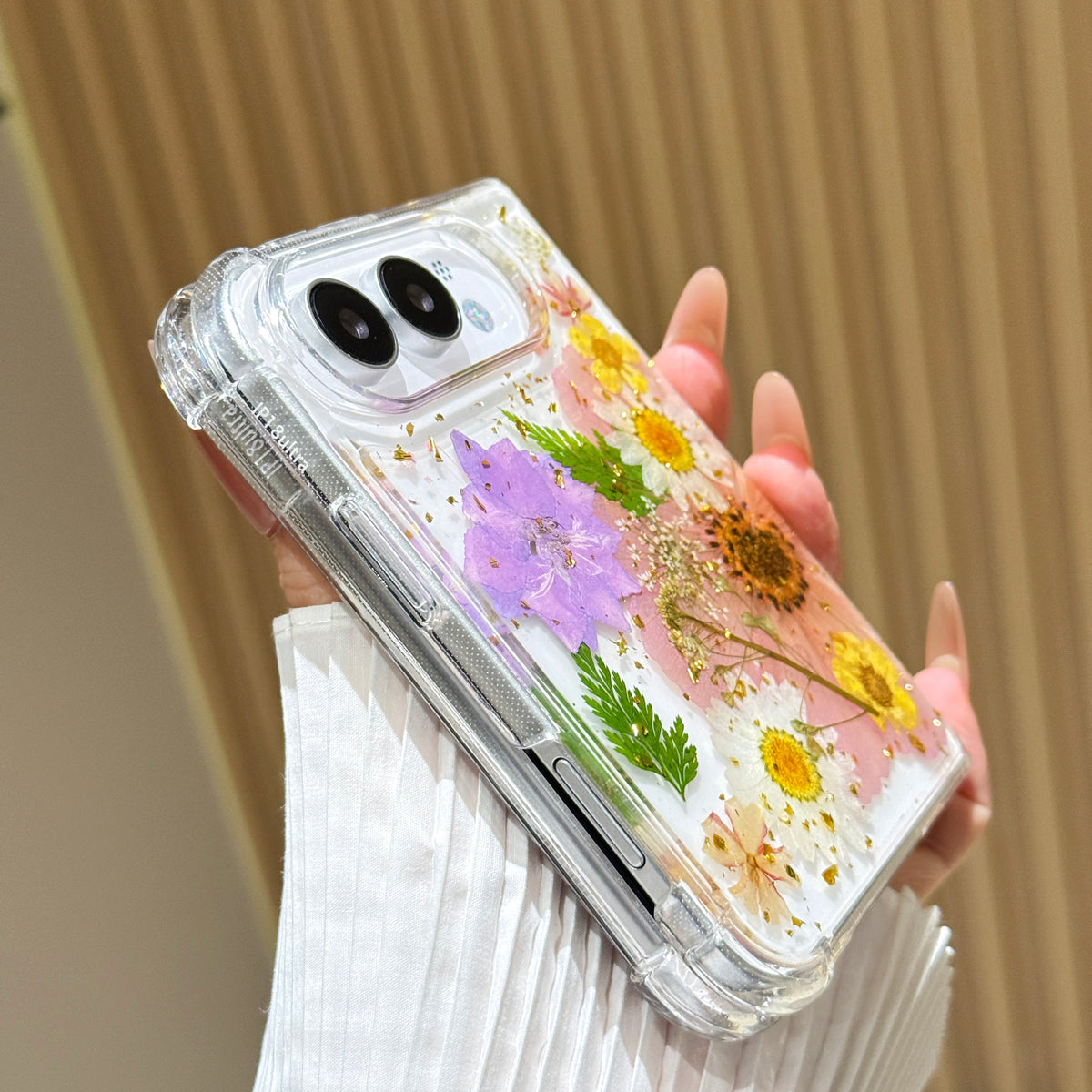 Side perspective view of a hand holding the clear iPhone DUO pressed flower case, displaying reinforced corner bumpers, precision side button alignment, and dual camera lens bezel.
