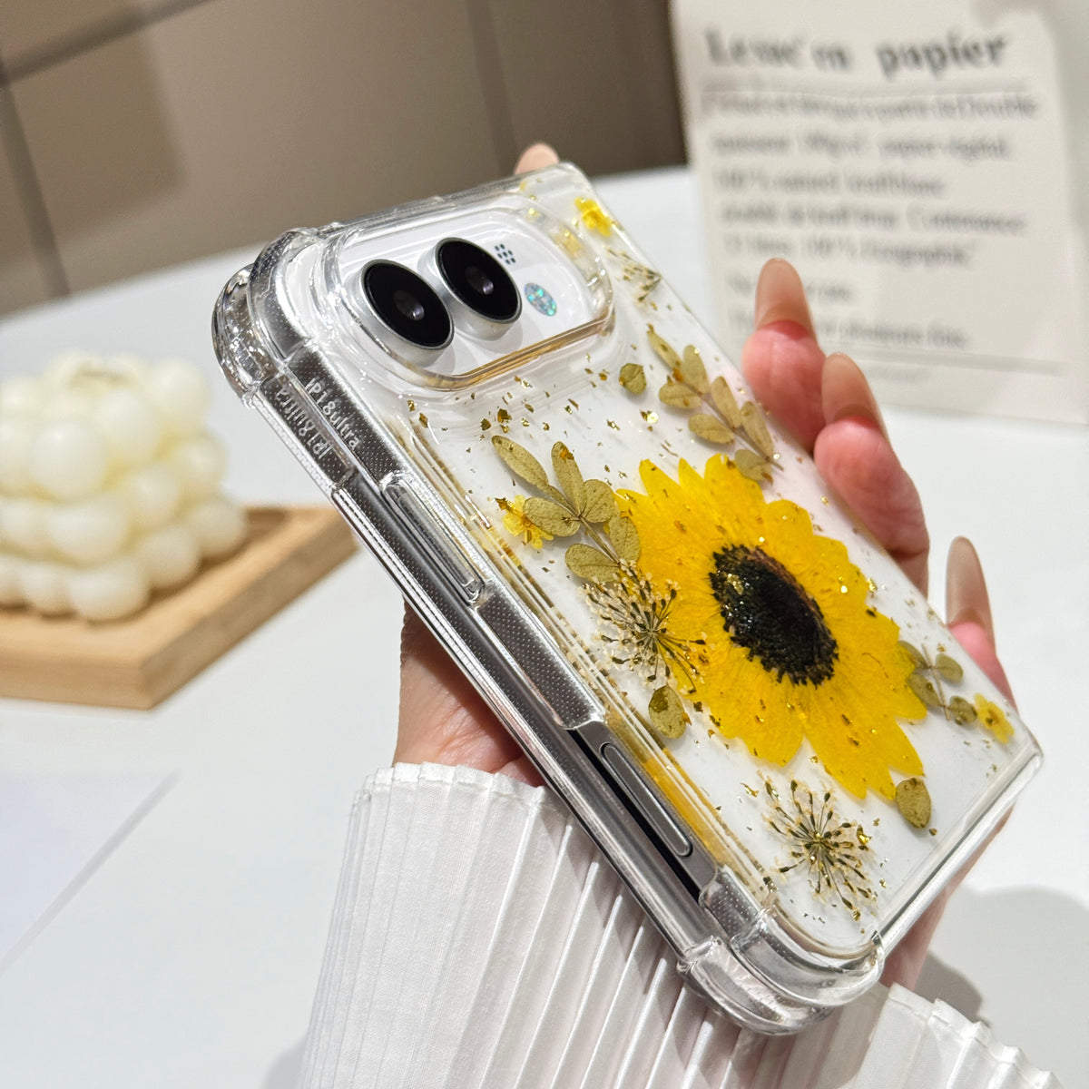Side perspective view of a hand holding the clear iPhone DUO pressed flower case, displaying reinforced corner bumpers, precision side button alignment, and dual camera lens bezel.