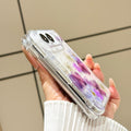 Side perspective view of a hand holding the clear iPhone DUO pressed flower case, displaying reinforced corner bumpers, precision side button alignment, and dual camera lens bezel.