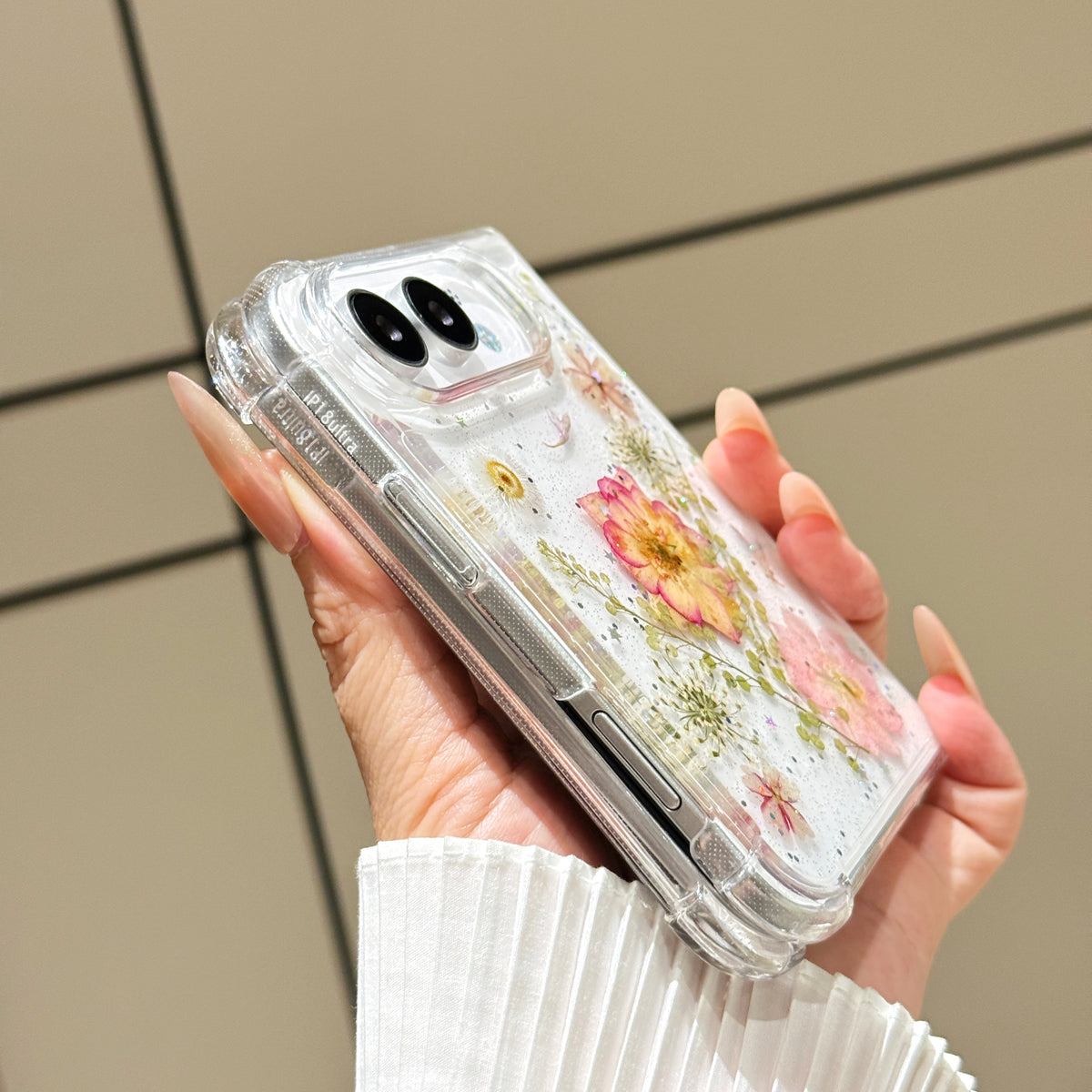 Side perspective view of a hand holding the clear iPhone DUO pressed flower case, displaying reinforced corner bumpers, precision side button alignment, and dual camera lens bezel.