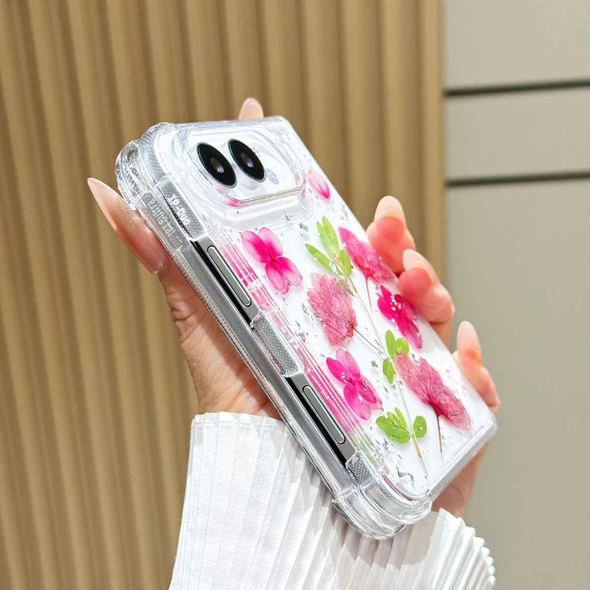 Side perspective view of a hand holding the clear iPhone DUO pressed pink flower case, displaying reinforced corner bumpers, precision side button alignment, and dual camera lens bezel.