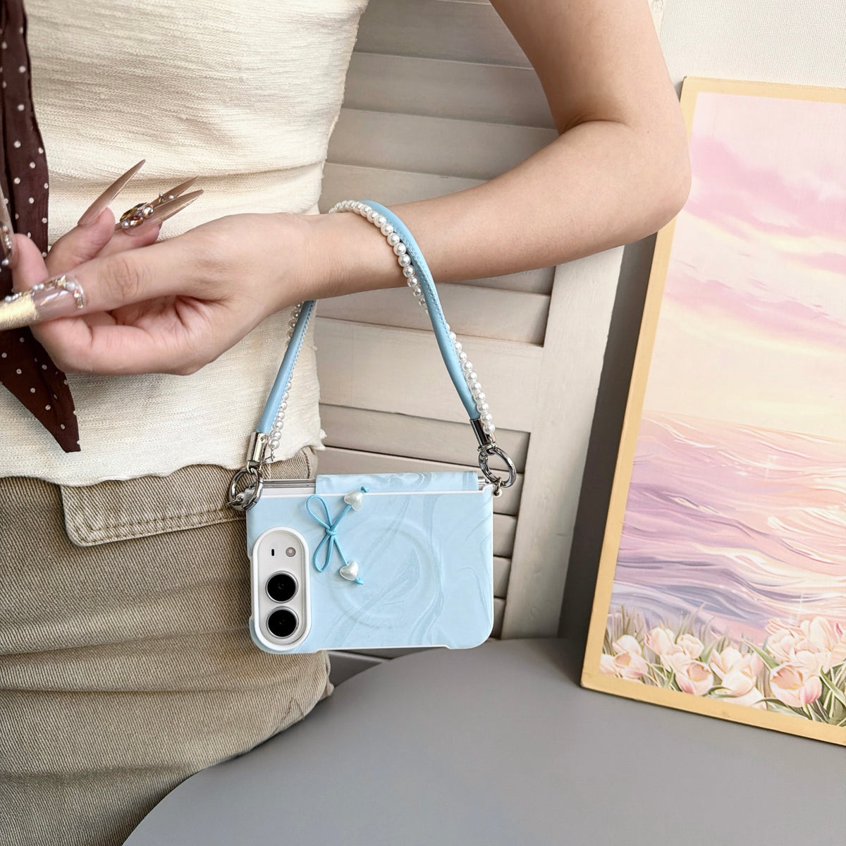 Sky blue marble swirl case for iPhone DUO held in hand displaying front lock screen display, 3D tied cord bow with pearl heart drops, raised dual-camera bezel, and attached blue leather pearl-lined lanyard wristlet strap.