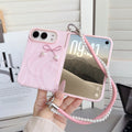 Soft blush pink marble swirl case for iPhone DUO held in hand displaying front lock screen display, 3D tied cord bow with pearl heart drops, raised dual-camera bezel, and attached pink leather pearl-lined lanyard wristlet strap.