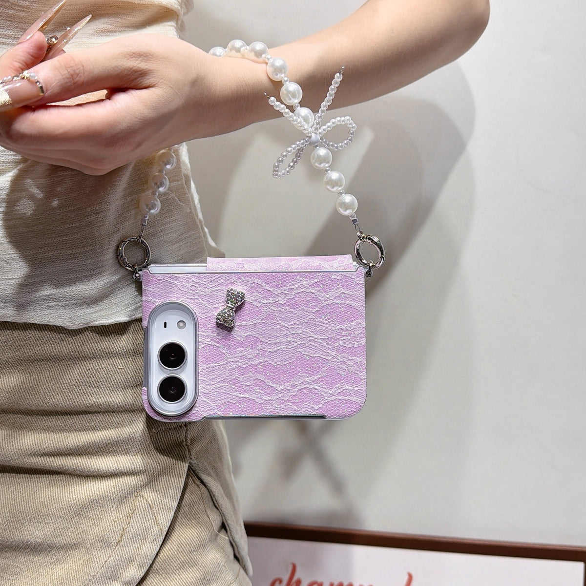 Soft lavender lace case for iPhone DUO held in hand showing front lock screen display, delicate glitter lace back texture, crystal bow charm, and attached pearl bow lanyard chain strap.