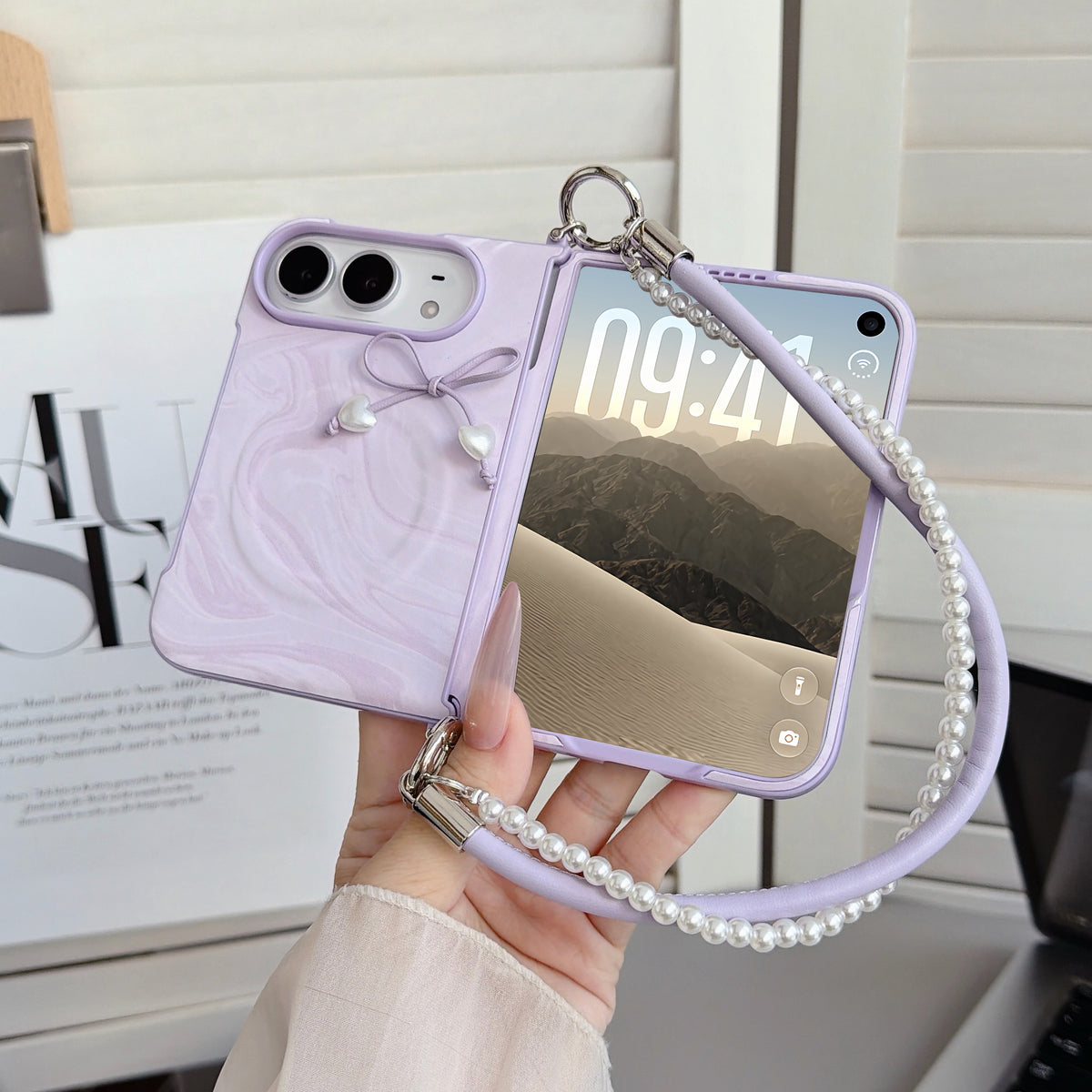 Soft pastel lavender purple marble swirl case for iPhone DUO held in hand displaying front lock screen display, 3D tied cord bow with pearl heart drops, raised dual-camera bezel, and attached purple leather pearl-lined lanyard wristlet strap.