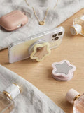 A star-shaped phone grip with a wooden star center and a clear outer shell, shown attached to a smartphone on a light wood surface with styled accessories including perfume bottles, a phone case, a necklace, and a small round mirror.