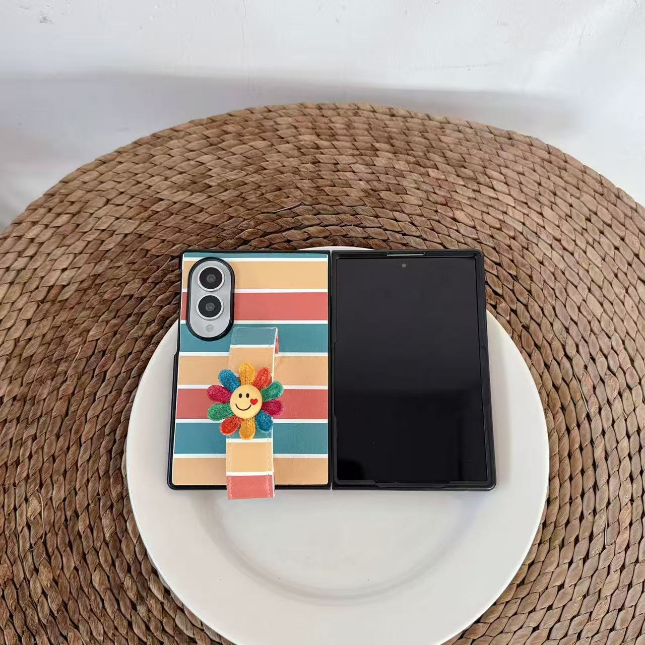 Top-down flat lay view of cute pastel rainbow striped Samsung Galaxy Z Fold case with smiley flower strap on a woven surface