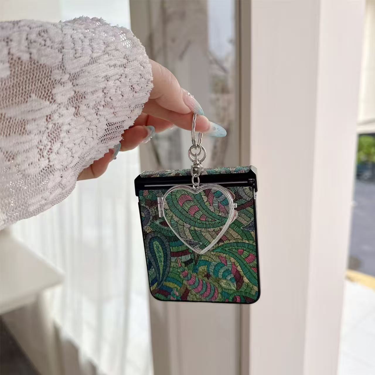 Woven emerald green paisley textured phone case for Samsung Galaxy Z Flip with hanging silver heart compact mirror held in hand.
