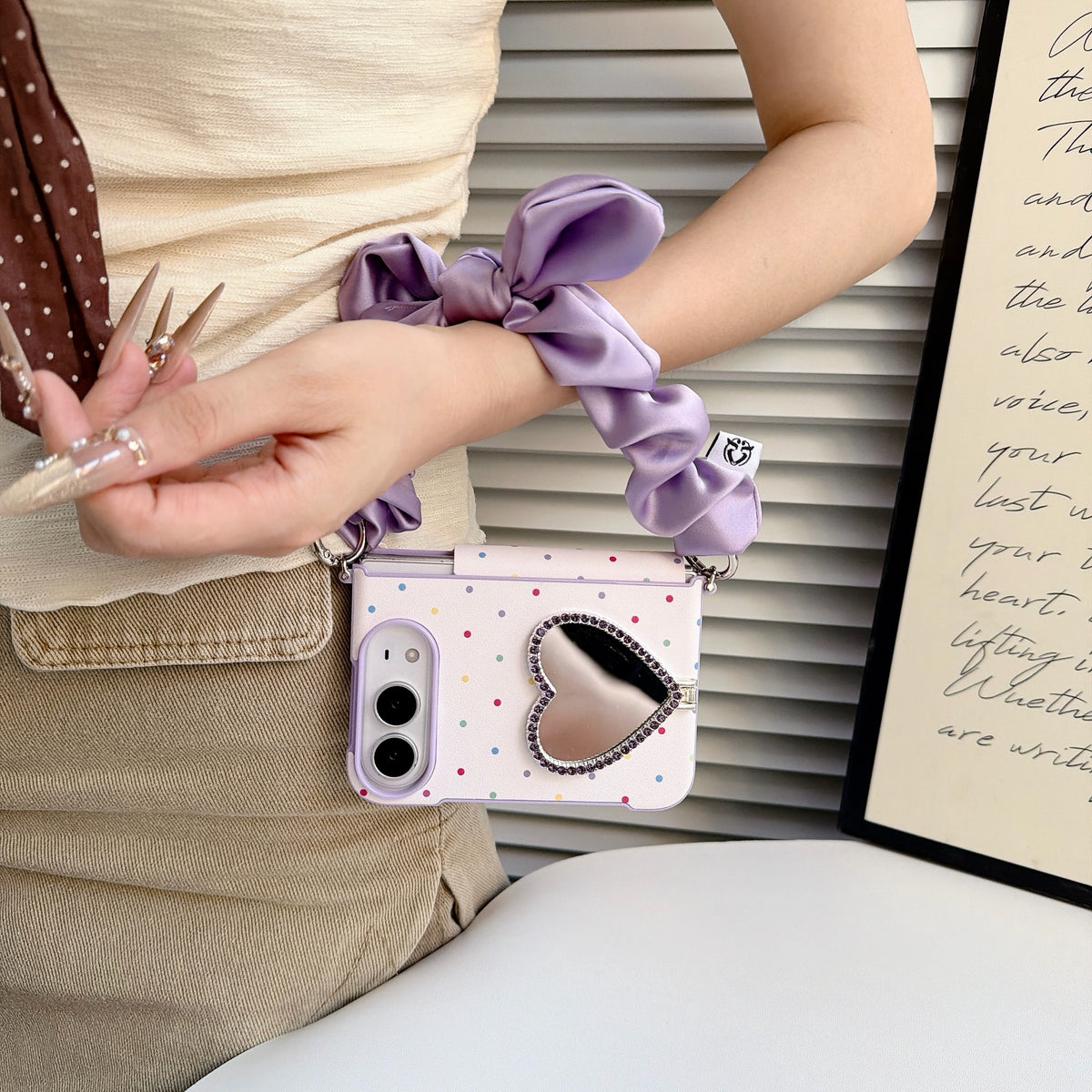 iPhone DUO polka dot case in lavender purple held by a model wearing the matching satin silk scrunchie wristlet strap, displaying the rhinestone heart mirror and dual-camera cutouts.
